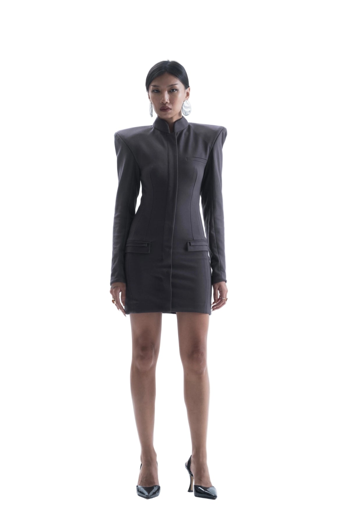 A model stands against a seamless white studio background wearing a dark gray structured mini dress with shoulder epaulets, a high mock neck, and front flap pockets, paired with black pointed-toe heels.