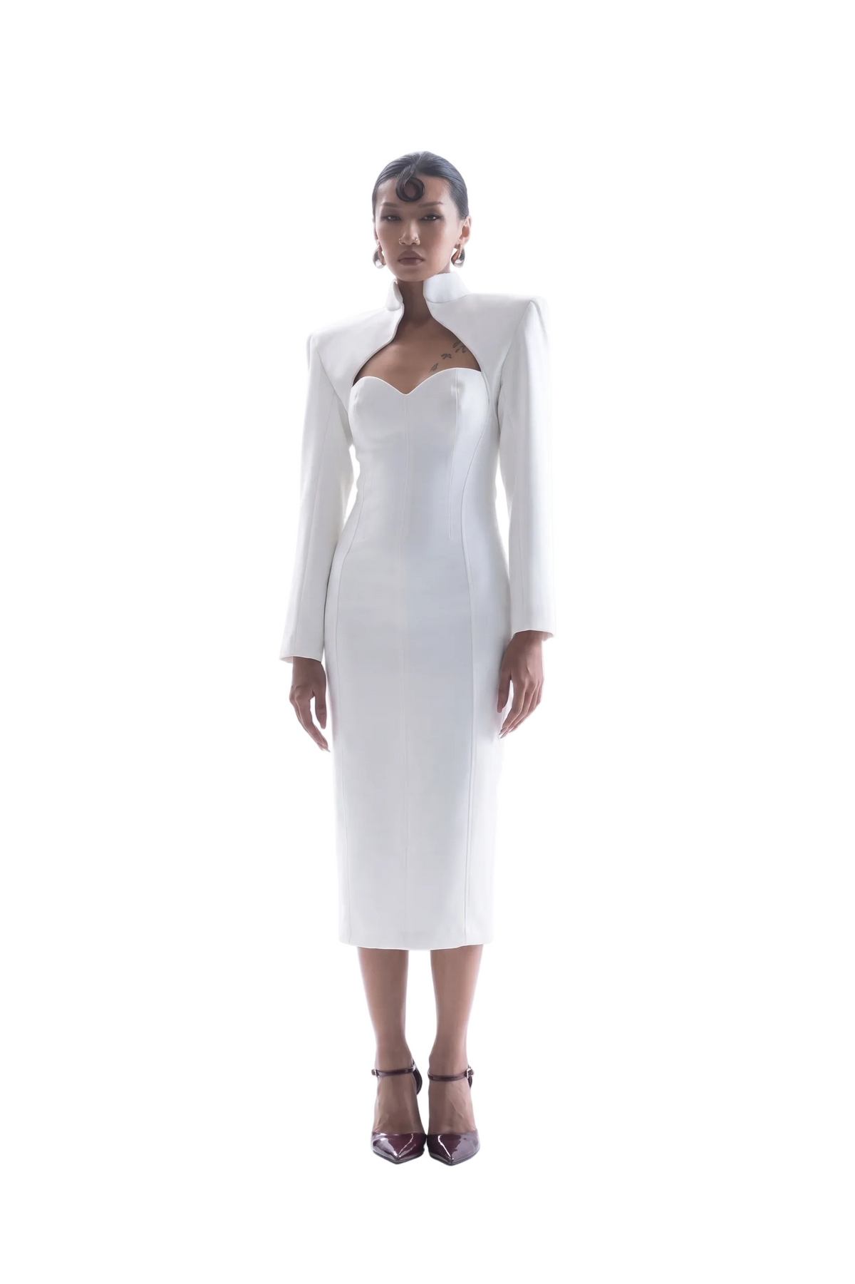 A model stands against a seamless white studio background wearing a white long-sleeve midi dress with structured shoulders, a sweetheart neckline, and a fitted silhouette, paired with dark pointed-toe heels.